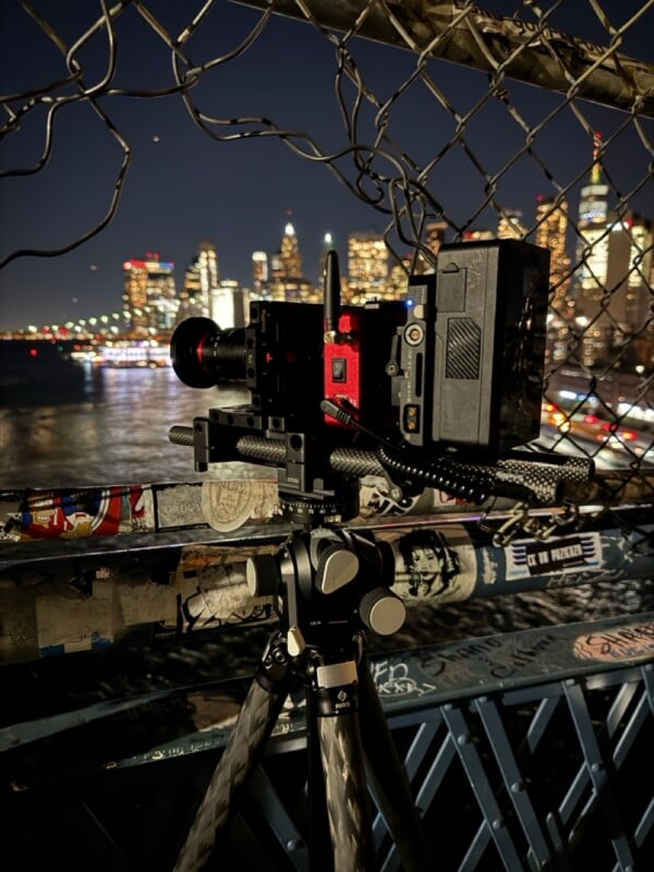 A professional cinema camera mounted on a tripod sits behind a chain-link fence overlooking a blurred city skyline at night.
