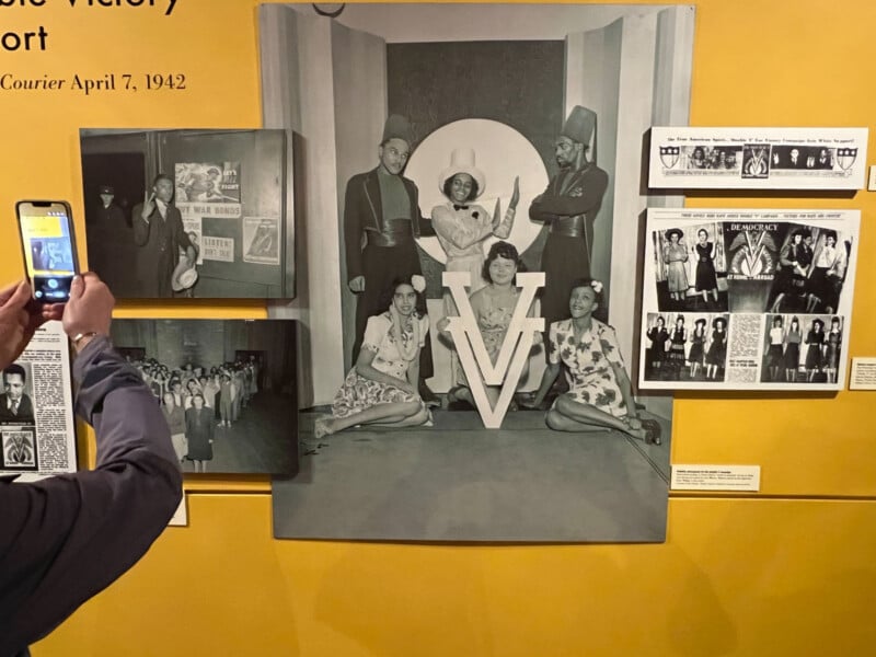 A museum display on a yellow wall features historical black-and-white photographs and newspaper clippings related to the "Double V" campaign, with a person's hand holding a smartphone to photograph the exhibit.