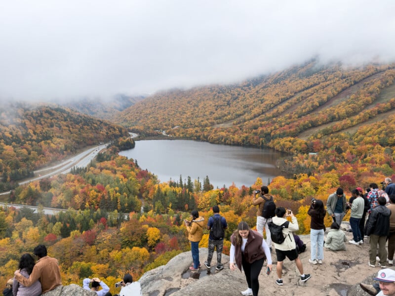 A group of people stand on a rocky overlook taking photos of a lake surrounded by autumn foliage and a highway, with mountains in the background.