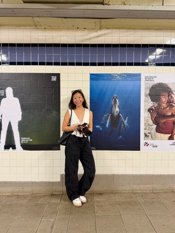A smiling woman with a camera around her neck stands in a subway station in front of three large posters on a tiled wall.