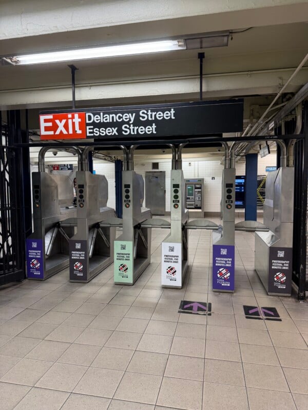 A row of subway turnstiles at the Delancey Street/Essex Street station, each featuring advertisements for a Fujifilm photography festival.