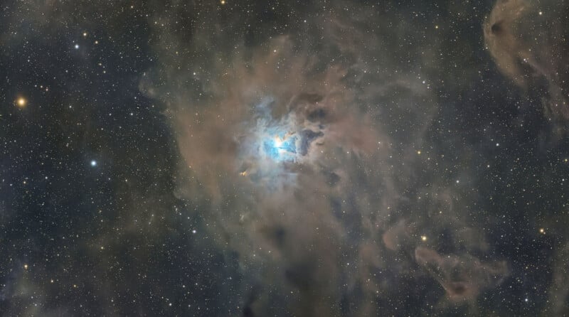 A bright blue reflection nebula surrounded by dark, wispy dust clouds against a dense field of stars.