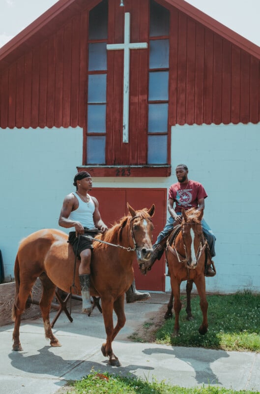 Two men sit on horseback in front of a white and red building with a large white cross and the number 223 above the door.