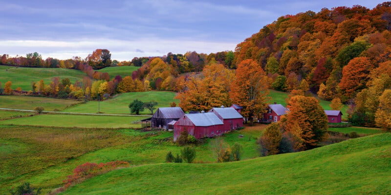 A rural landscape in autumn features red barns and a farmhouse nestled among rolling green hills and vibrant, colorful trees under a cloudy sky.