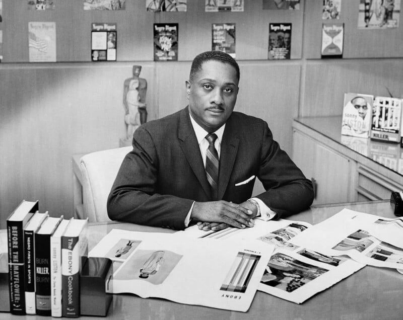 John H. Johnson sits at a desk in a suit and tie, with books and magazine proofs spread before him and shelves of publications in the background.