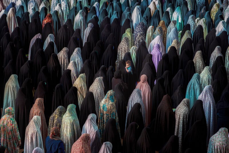 A large crowd of women wearing various styles of headscarves and full-body coverings stands together, with a small child visible in the center.