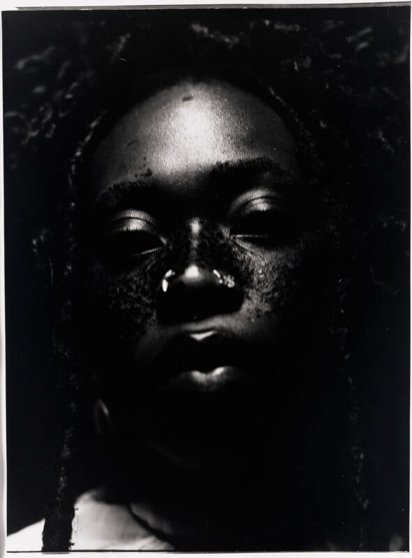 A close-up, high-contrast black and white portrait of a person with dark skin, visible freckles, and braided hair.