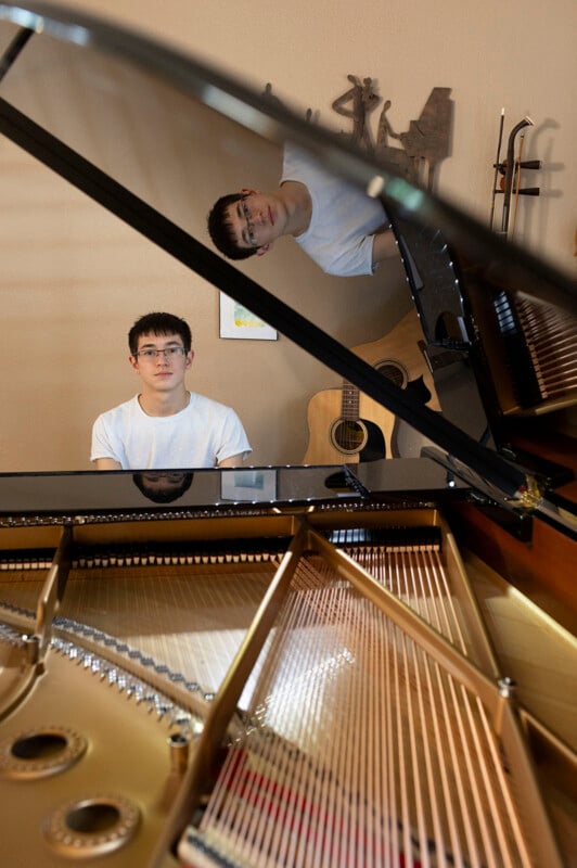 A young man with glasses and a white t-shirt sits at a grand piano, his reflection visible in the instrument's polished lid.