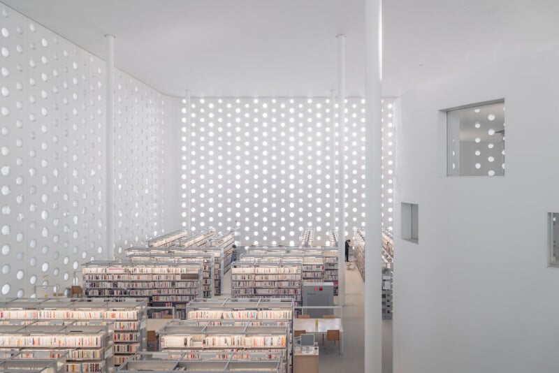 A bright, minimalist library interior features rows of bookshelves under a high ceiling with walls patterned by circular cutouts.