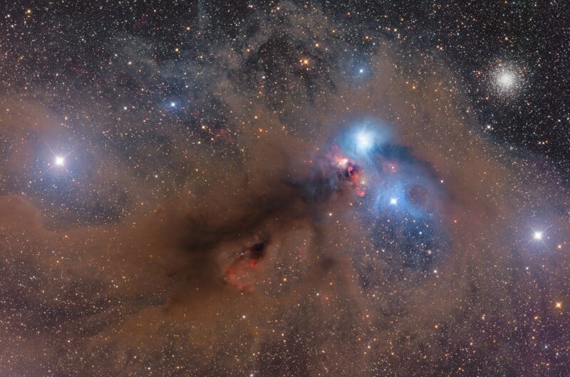 A vibrant nebula featuring glowing blue reflection clouds, dark dust lanes, and a dense cluster of stars in the upper right.
