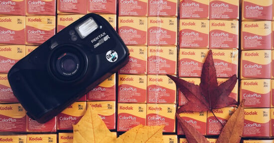 A black Pentax Zoom 90-WR camera rests on a grid of Kodak ColorPlus 200 film boxes, accented by several dried autumn leaves.
