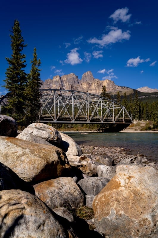 A metal truss bridge spans a river in front of a jagged mountain range under a clear blue sky with scattered clouds.