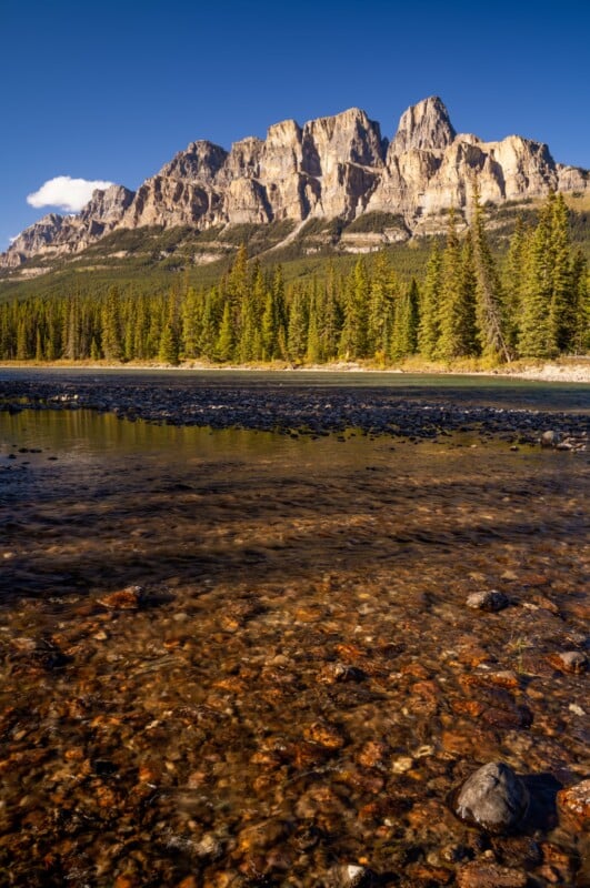 A clear, shallow river flows over a rocky bed toward a dense evergreen forest beneath a towering, rugged mountain range.
