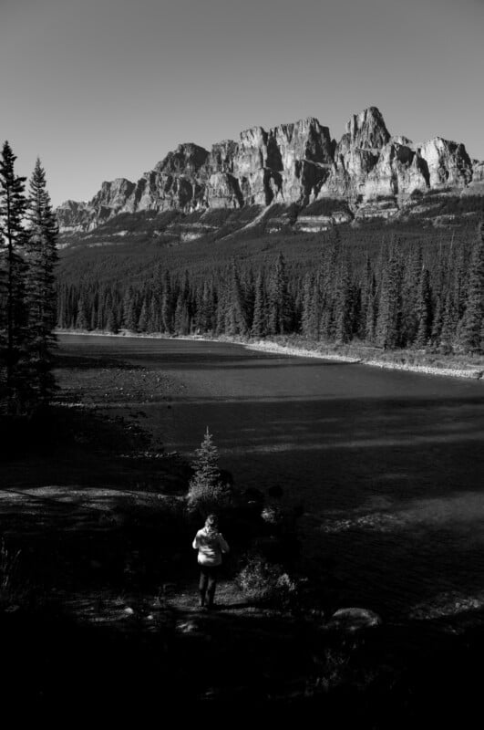 A person stands on a riverbank looking toward a large, rugged mountain range rising above a dense pine forest.