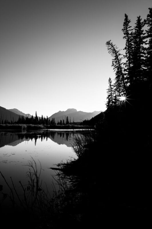 A black and white landscape shows a calm lake reflecting distant mountains, with a sunburst shining through silhouetted trees.