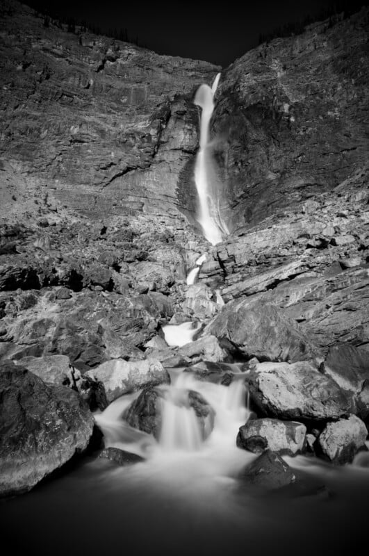 A long-exposure black and white shot of a tall waterfall cascading down a rocky mountain cliff into a stream below.