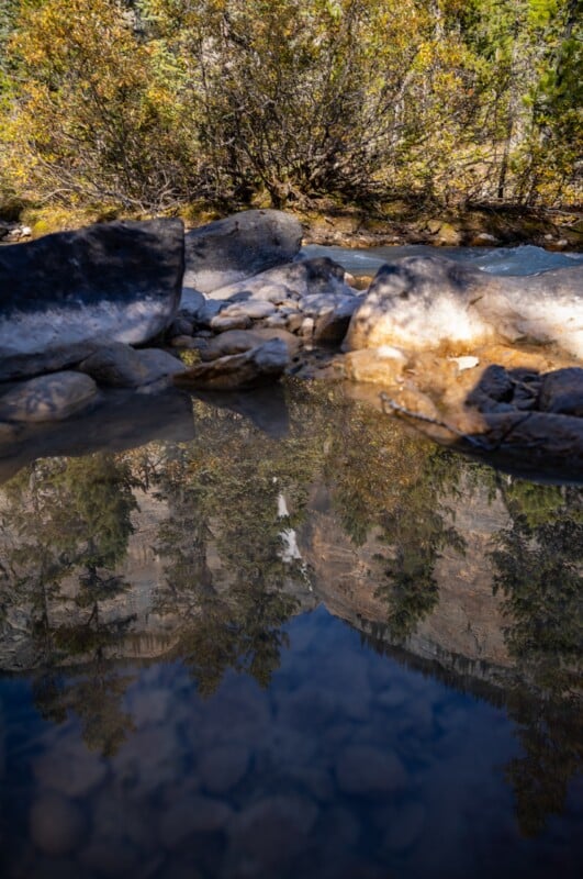 A clear pool of water in the foreground reflects a mountain slope, trees, and a waterfall, with a flowing stream and rocks behind.