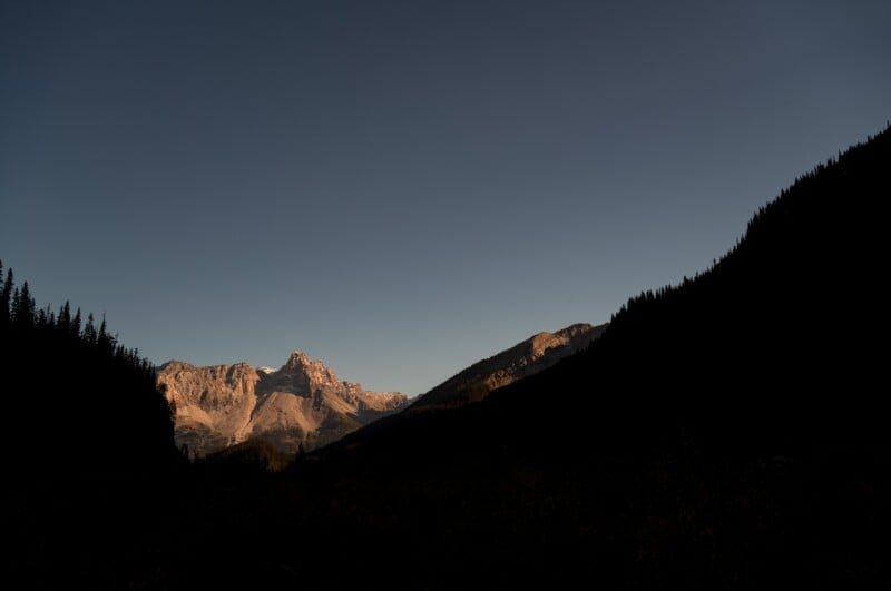 Sunlight illuminates the peaks of a mountain range, while the surrounding valley and slopes remain in deep shadow.