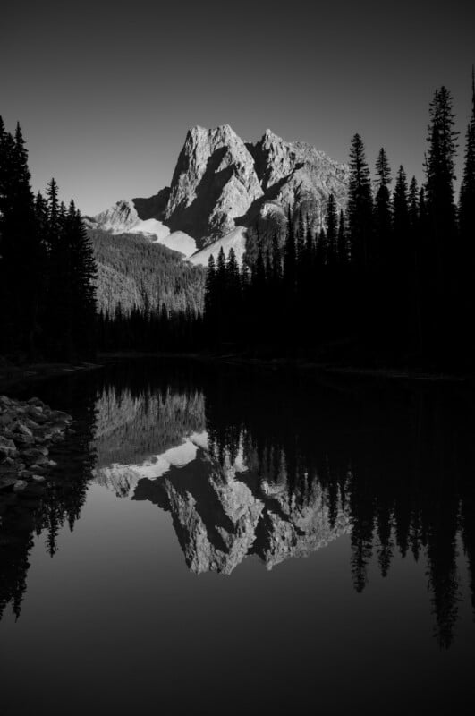 A black and white landscape shows a jagged mountain peak reflecting perfectly in the still, dark water of a lake, framed by silhouettes of pine trees.