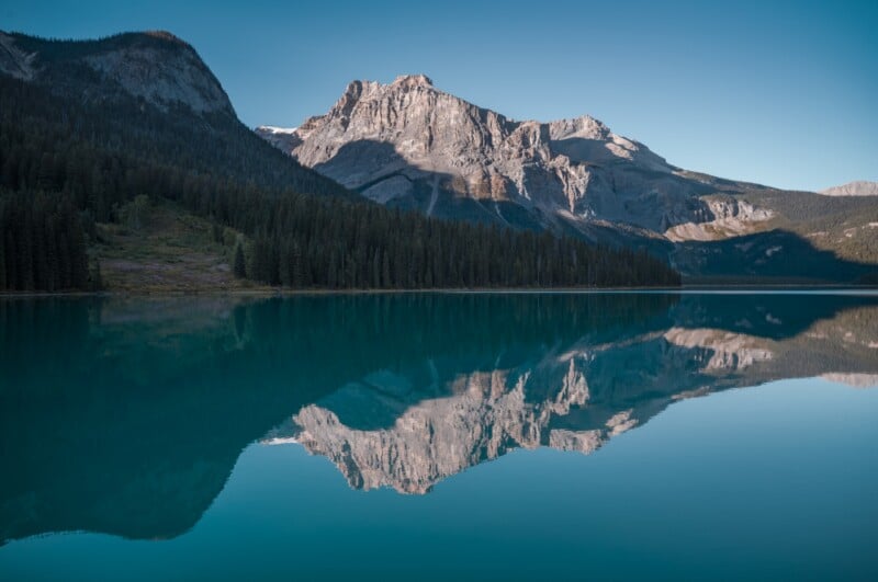 A jagged mountain peak with a partial shadow is reflected in the calm, turquoise water of a lake, bordered by a dense forest.