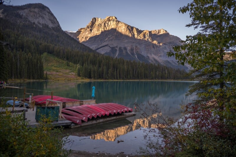 A wooden dock holds a stack of red canoes on a calm lake, with a large mountain reflecting in the water under a clear sky.