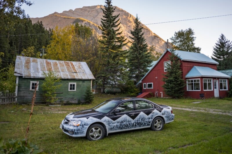 A black sedan with a white mountain-themed paint job is parked on a grassy lawn in front of a green shed and a red house, with a large mountain in the background.