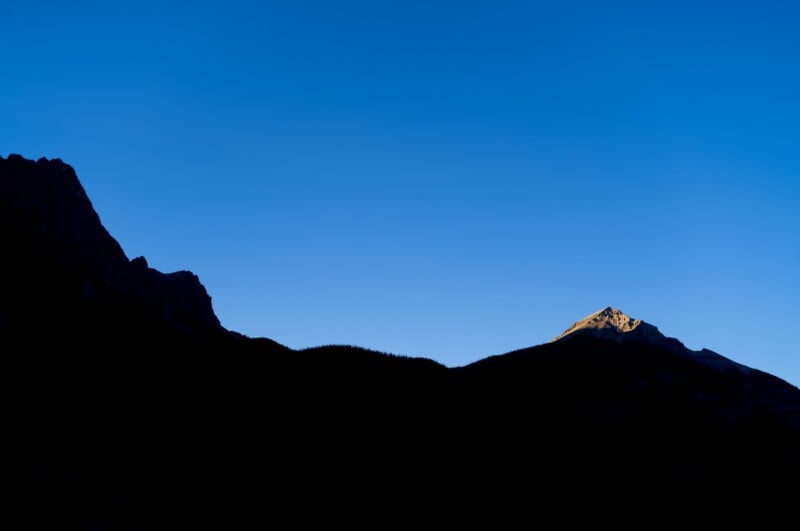 A silhouette of a mountain range against a clear blue sky, with the peak of one mountain illuminated by golden sunlight.