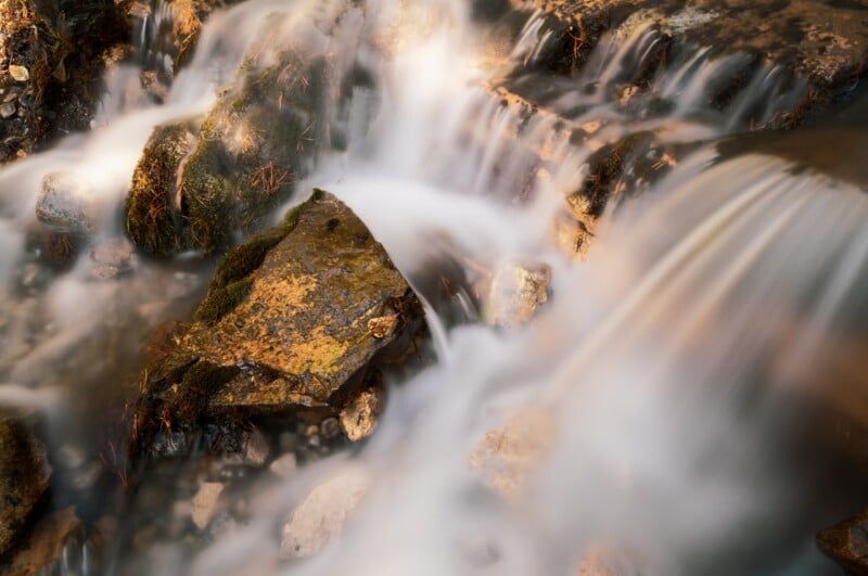 A long-exposure shot of a small stream flowing over mossy, textured rocks and pebbles.