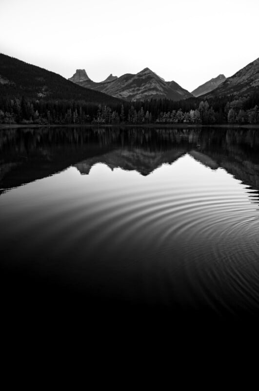 A black and white landscape shows a calm lake reflecting a mountain range and a forest, with concentric ripples in the water.
