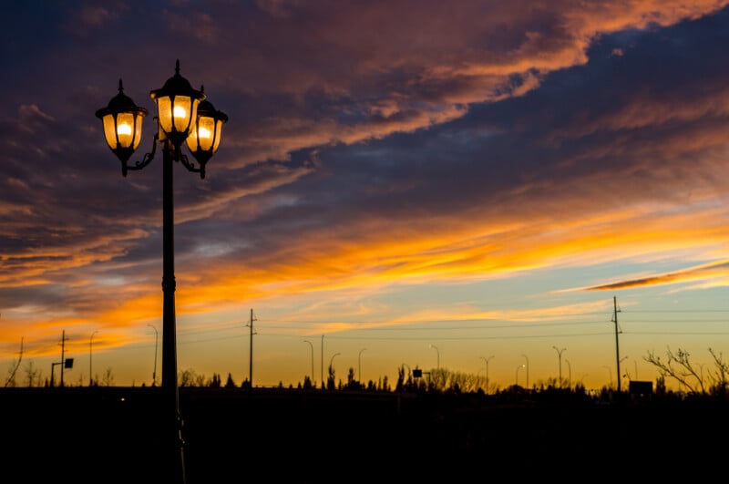 A lit three-headed street lamp stands in the foreground against a vibrant orange and purple sunset sky over a silhouetted landscape.
