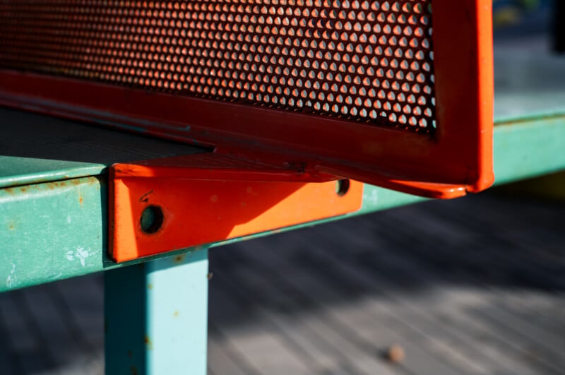 A close-up view of an orange metal bracket connecting a perforated orange backrest to a teal metal bench frame.