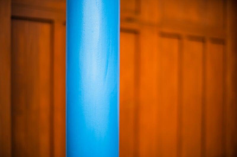 A bright blue, cylindrical pillar stands vertically in the foreground against a blurred background of wooden wall paneling.