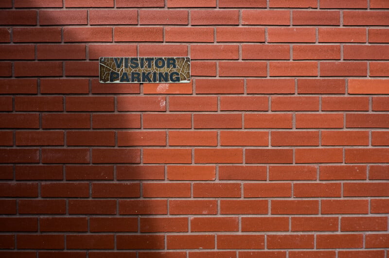A weathered "VISITOR PARKING" sign is mounted on a red brick wall, partially cast in shadow.
