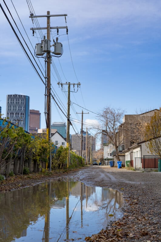A gravel alleyway with a large puddle in the foreground reflects a row of utility poles and a city skyline under a blue sky.