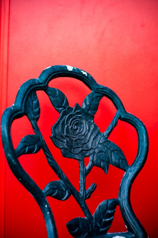 A close-up of a dark, ornate metal chair back featuring a rose motif, set against a vibrant, solid red background.