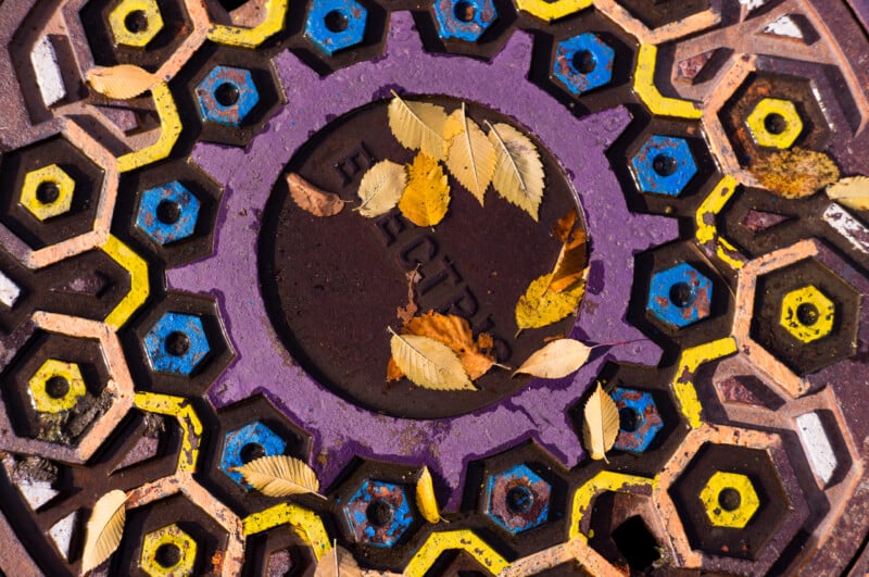 A painted metal utility cover with a geometric pattern of yellow and blue hexagons, featuring a central purple ring and scattered fallen autumn leaves.
