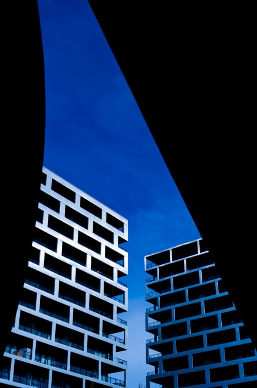 Two modern, grid-patterned apartment buildings stand against a deep blue sky, framed by dark, abstract silhouettes on the sides.