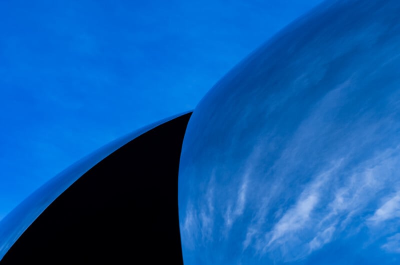 A smooth, curved metallic surface reflects a blue sky with wispy clouds, creating a sharp contrast against a dark shadow.