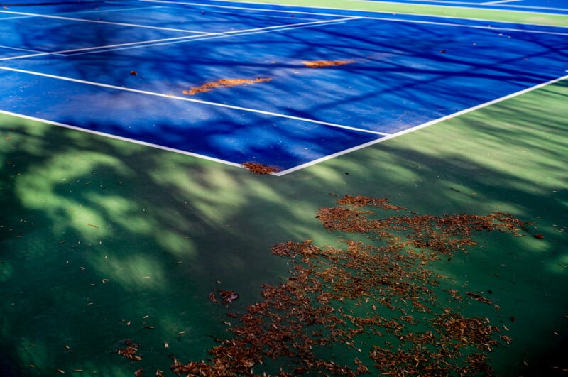 A high-angle view of a tennis court with blue and green surfaces, scattered with dry leaves and tree shadows.