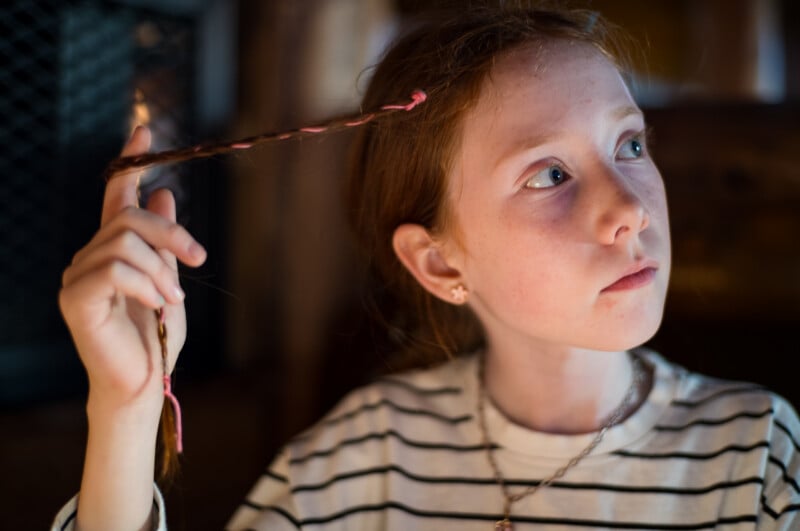 A young girl with red hair and freckles looks upward while holding a thin, pink-threaded braid near her face.