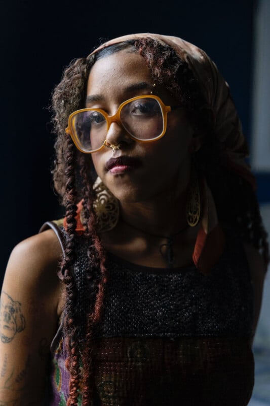 A person with braided hair and a headscarf wears large, yellow-rimmed glasses, a septum ring, and dangling earrings.