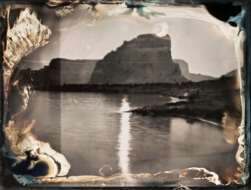A wet plate collodion photograph shows a large, dark rock formation rising above a calm body of water with a bright reflection of light on the surface.