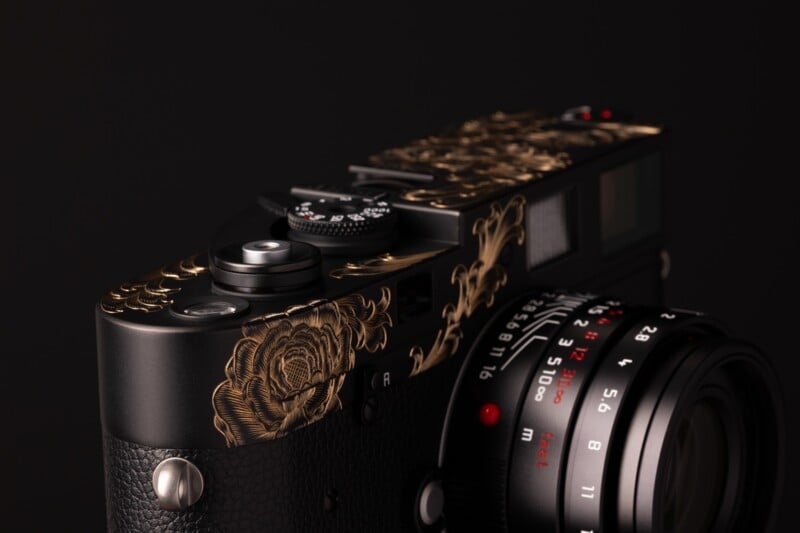 A close-up view of a black camera body featuring intricate gold floral engravings on its top plate and a lens with red markings.