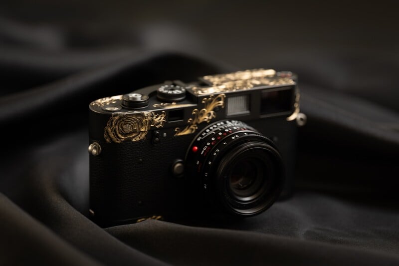 A black rangefinder-style camera with intricate gold floral engravings on its top plate, resting on dark, draped fabric.
