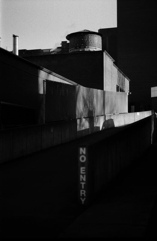 A black and white view of a concrete ramp with a vertical "NO ENTRY" sign, leading toward a brick building with a water tower.