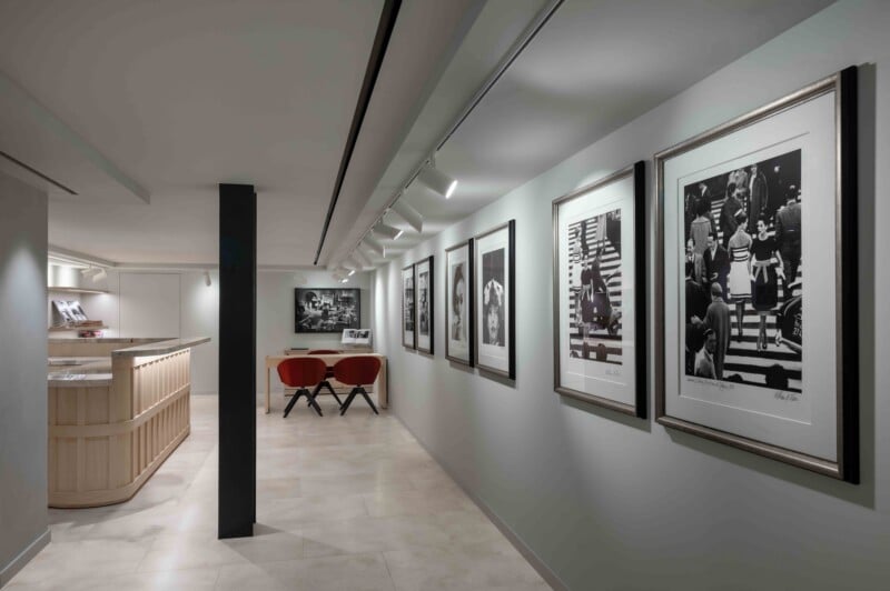 A modern interior features a light-colored hallway lined with framed black-and-white photographs, a wooden reception desk, and two red chairs at a small table.