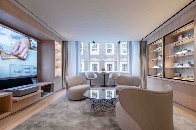 A modern lounge area features three beige armchairs arranged around a glass-topped coffee table on a grey rug, with a large television, display cabinets, and a window overlooking a brick building.