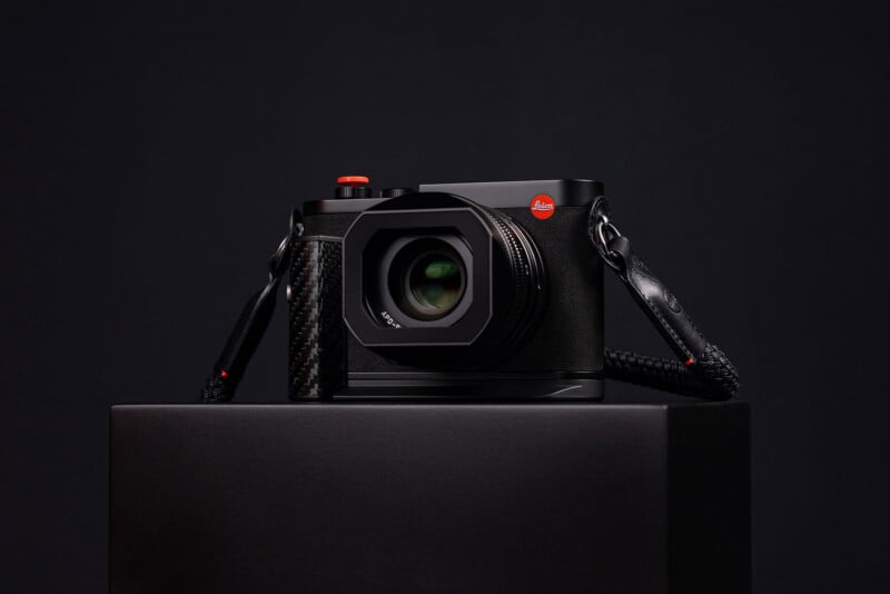 A black Leica camera with a carbon fiber grip and an orange shutter button sits on a black pedestal against a dark background.