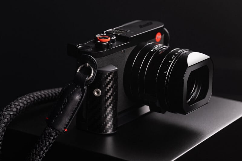 A black digital camera with a carbon fiber grip and a red shutter button sits on a dark surface, attached to a braided black strap.