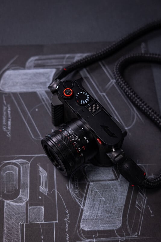 A black digital camera with a red shutter button and a woven strap rests on a dark surface featuring a white technical sketch of a vehicle.